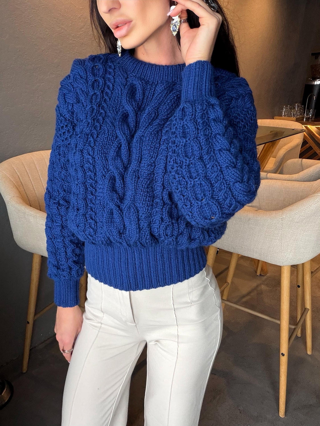 May include: A vibrant blue cable-knit sweater with a crew neck and long sleeves. The sweater features a textured pattern and a ribbed hem. Paired with white pants, the sweater is a stylish addition to any wardrobe.
