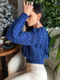 May include: A cobalt blue cable-knit sweater with a crew neck and long sleeves. The sweater features a textured pattern and a ribbed hem. The sleeves have a puffed design. The person is wearing beige pants.