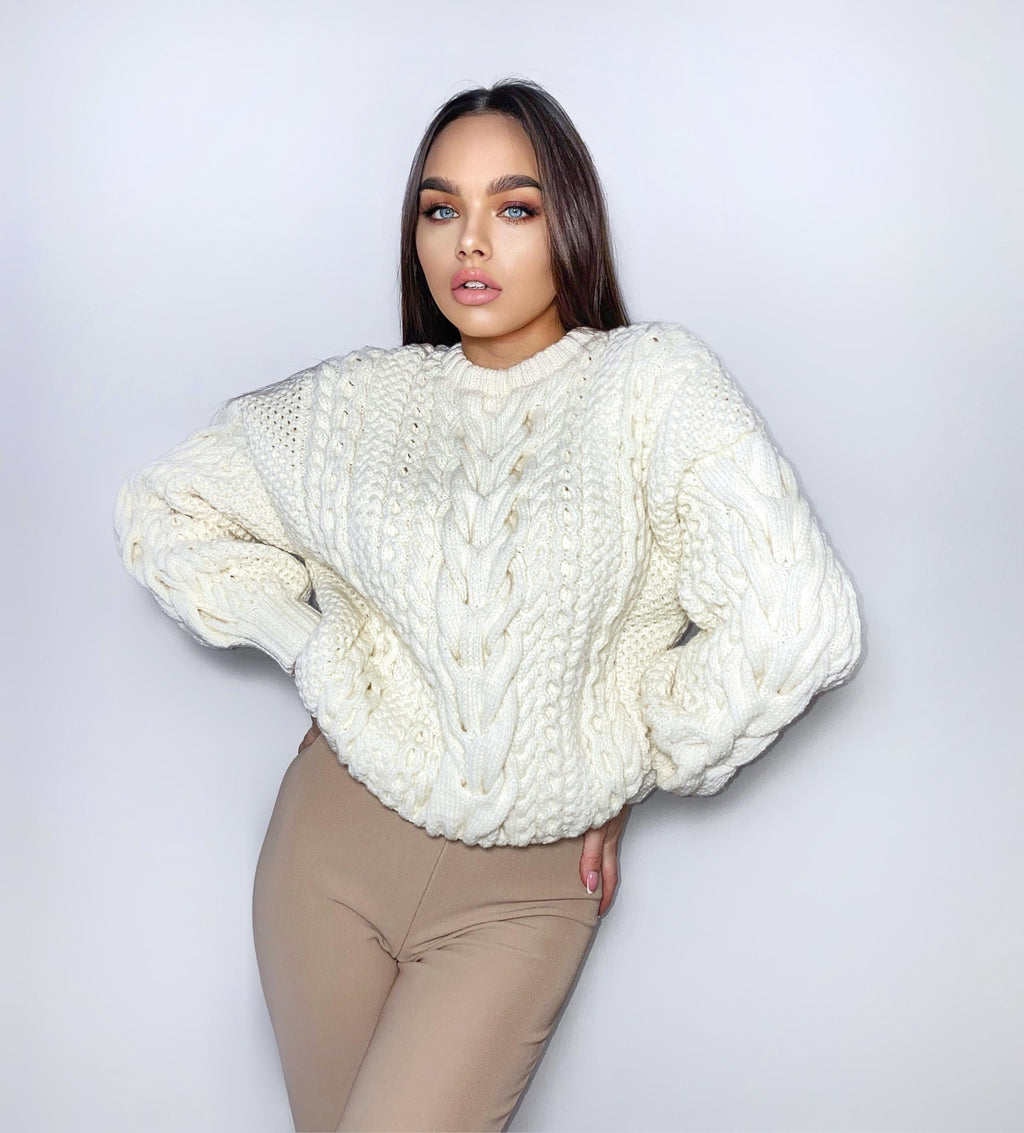 May include: A white cable knit sweater with a chunky texture. The sweater has a round neckline and long sleeves.