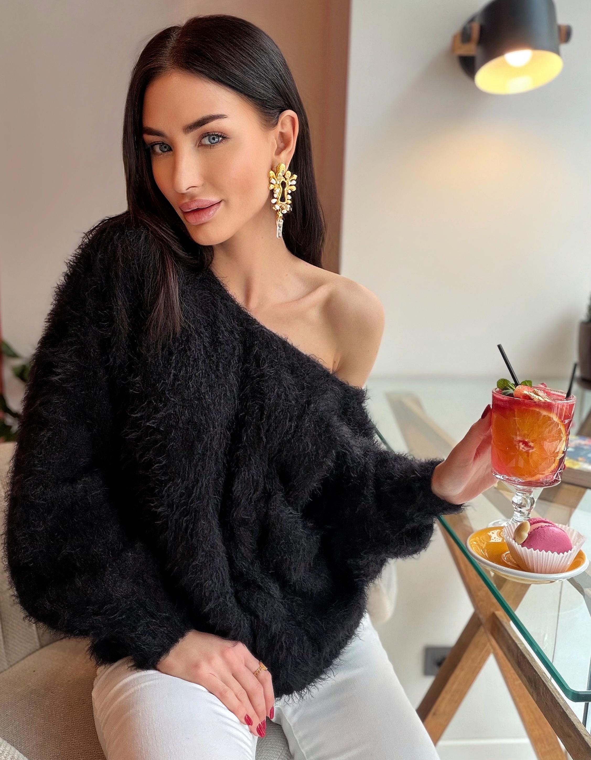 May include: A black, fluffy, off-the-shoulder sweater paired with white pants. The image also features gold earrings and a cocktail with fruit slices. The setting appears to be a cafe or restaurant.