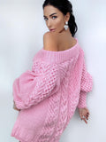 May include: A light pink, off-the-shoulder sweater with a cable knit design. The sweater has long sleeves with a puffy, textured design. The model is wearing silver heart-shaped earrings and rings.