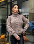 May include: A taupe cable-knit turtleneck sweater with intricate patterns. The sweater features a high, ribbed collar and long sleeves. The design includes a mix of cable and textured knit stitches. The model is wearing black pants.