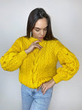 May include: A yellow cable knit sweater with a round neckline and long sleeves. The sweater has a loose fit and a textured pattern.