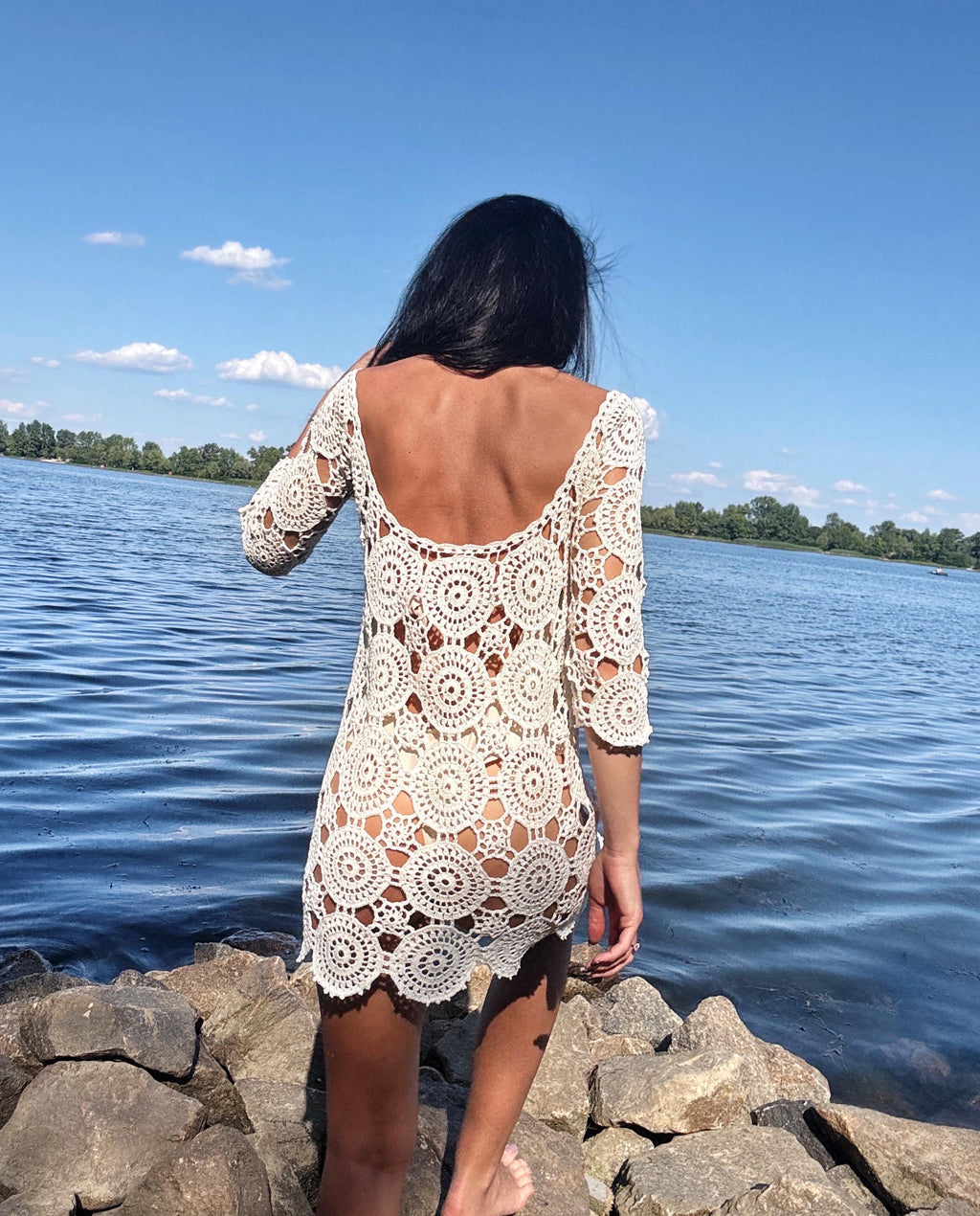 May include: A cream-colored crochet dress with a round neckline and three-quarter sleeves. The dress features a circular pattern design and is worn outdoors near a body of water.