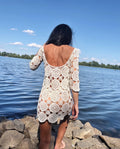 May include: A cream-colored crochet dress with a round neckline and three-quarter sleeves. The dress features a circular pattern design and is worn outdoors near a body of water.