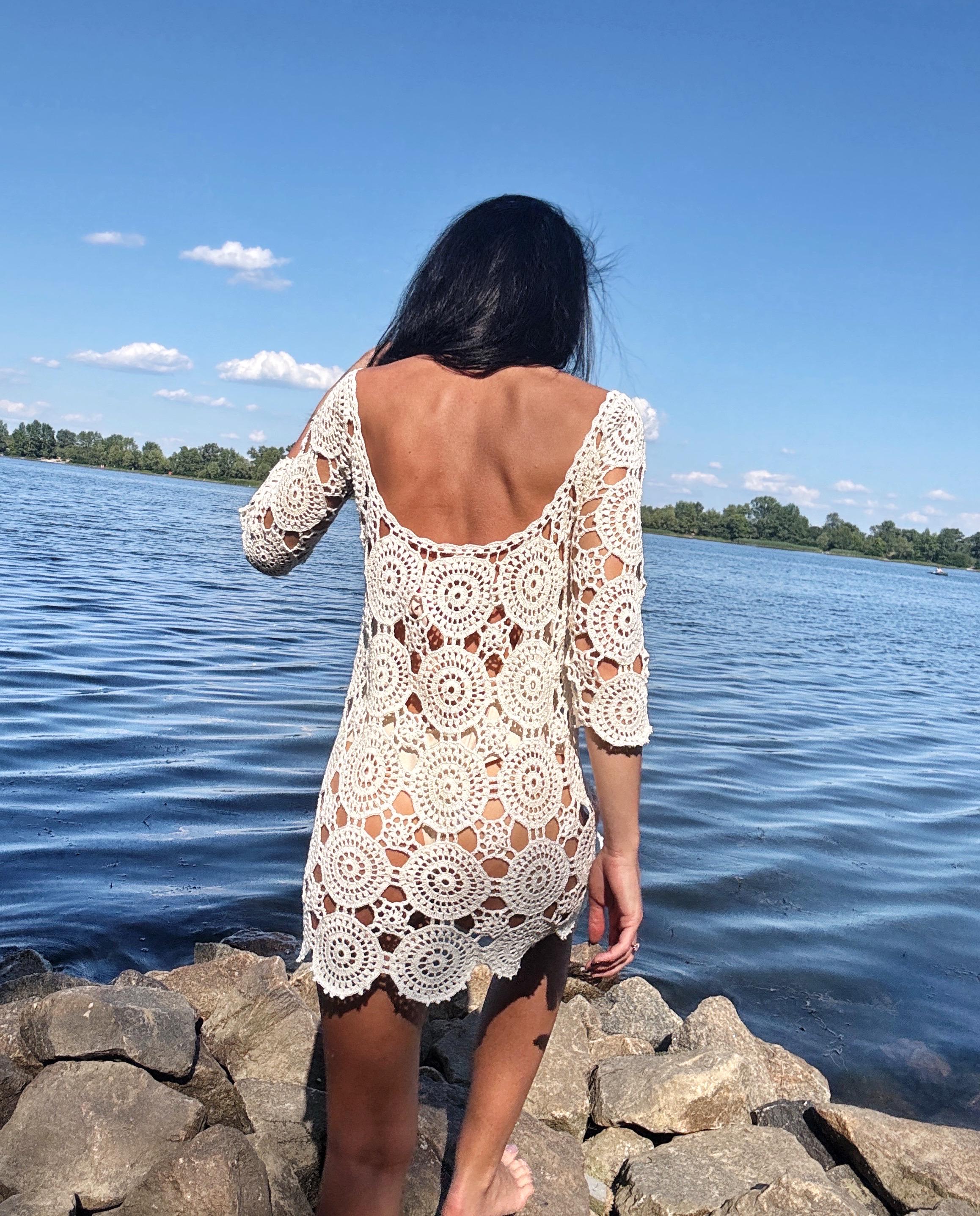 May include: A cream-colored crochet dress with a round neckline and three-quarter sleeves. The dress features a circular pattern design and is worn outdoors near a body of water.