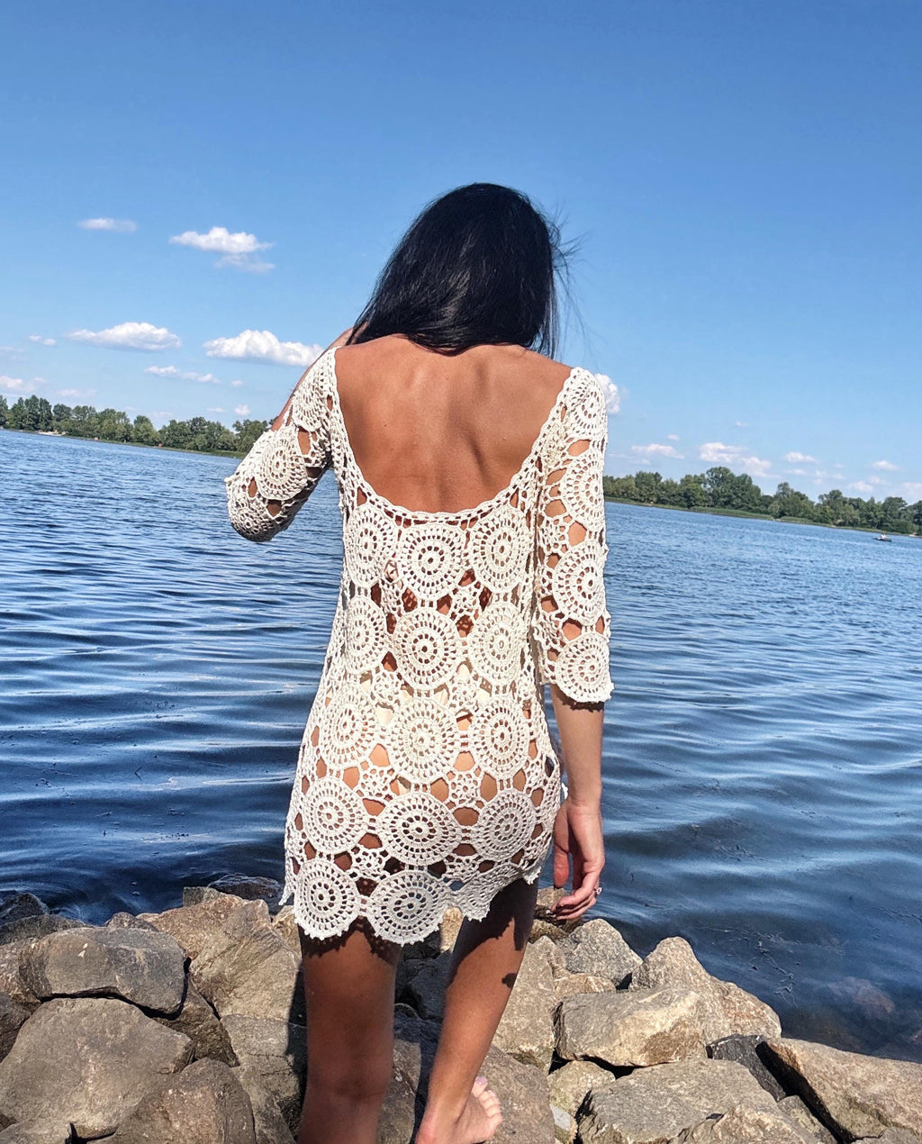 May include: A cream-colored crochet dress with a round neckline and three-quarter sleeves. The dress features a circular pattern design and is worn outdoors near a body of water.