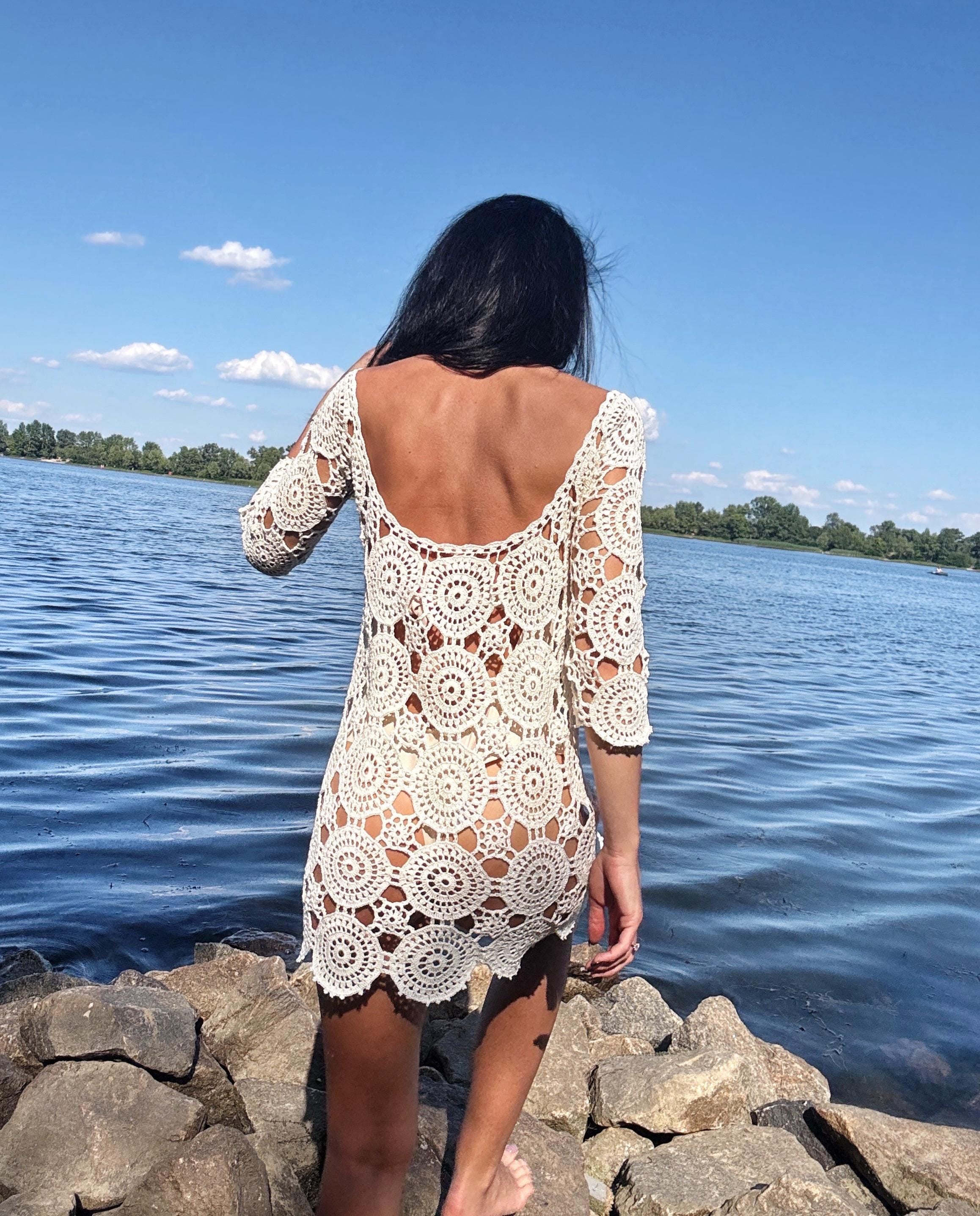 May include: A cream-colored crochet dress with a round neckline and three-quarter sleeves. The dress features a circular pattern design and is worn outdoors near a body of water.
