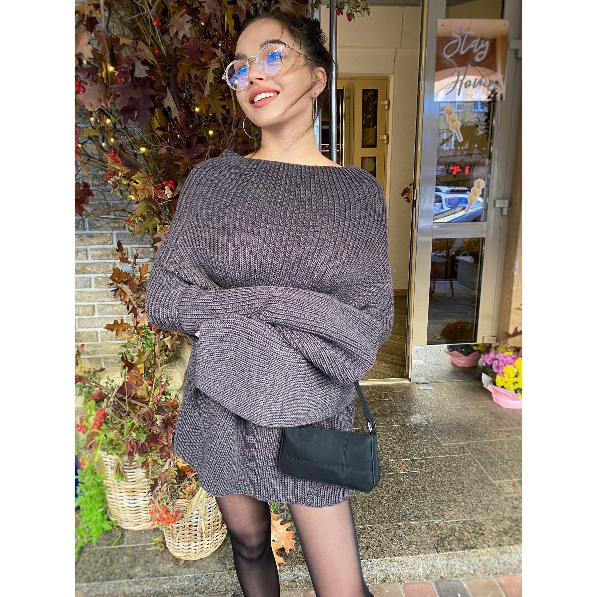 May include: A woman wearing a dark gray knitted sweater dress with a black shoulder bag. The sweater has a loose fit and long sleeves. The woman is standing in front of a building with a sign that says "Stay Home".