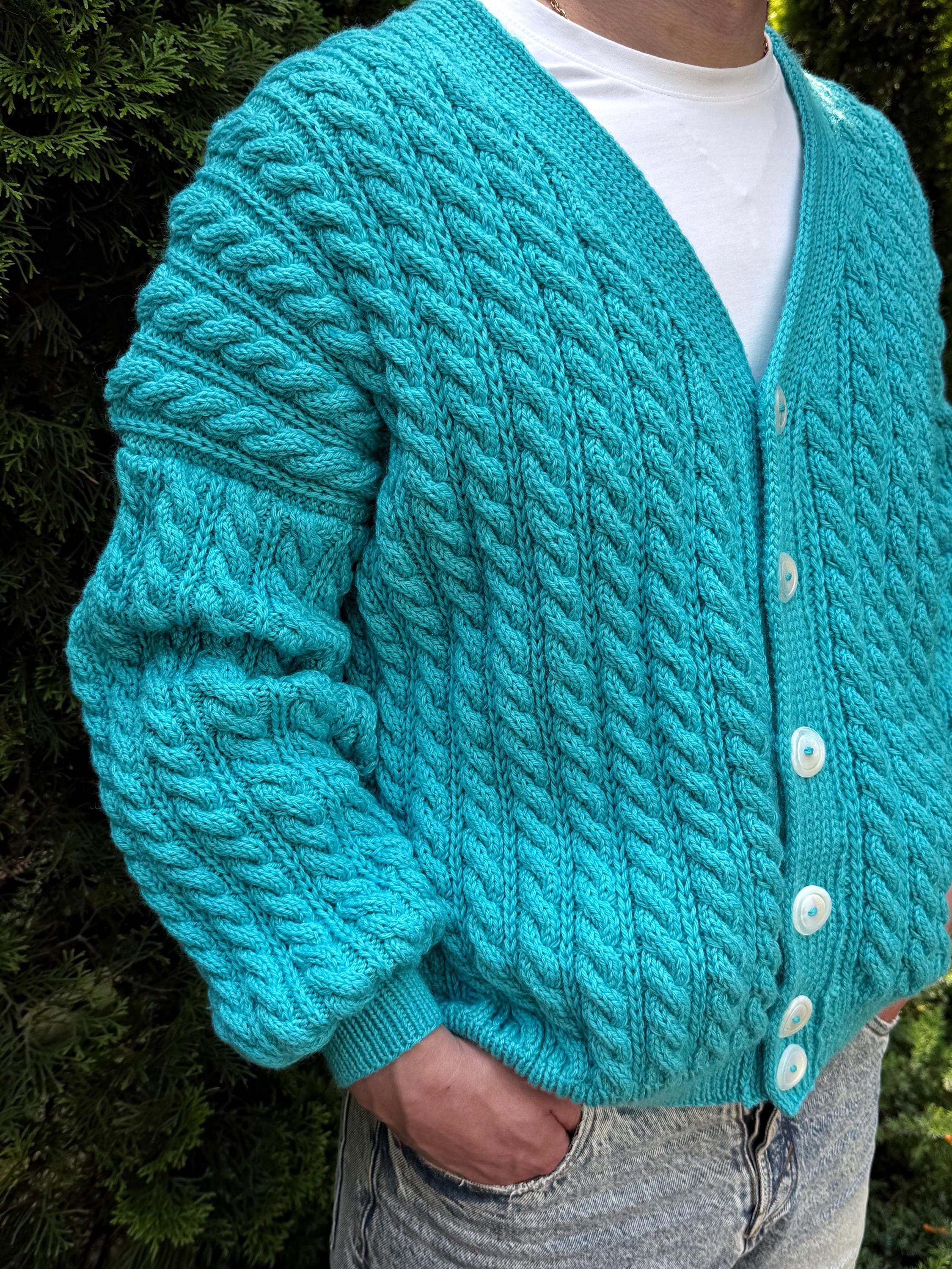 May include: A turquoise cable-knit cardigan with a V-neck and white buttons. The chunky knit sweater has a textured pattern and long sleeves. The person is wearing a white t-shirt underneath and blue jeans.