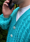 May include: A close-up of a turquoise cable-knit cardigan with two large white buttons. The cardigan is worn over a white t-shirt. The sleeves have a textured pattern. The person is holding a purple phone to their ear.