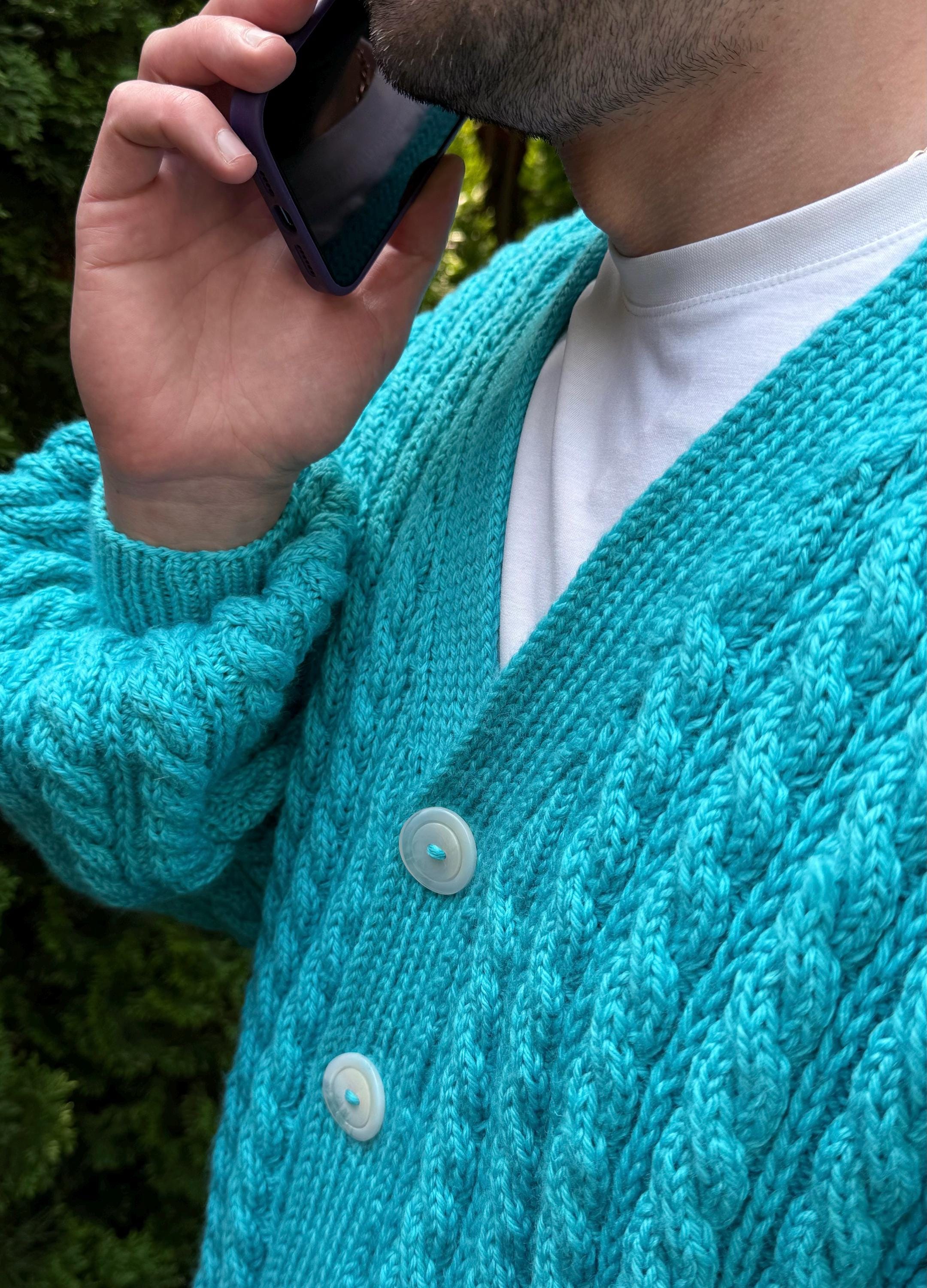 May include: A close-up of a turquoise cable-knit cardigan with two large white buttons. The cardigan is worn over a white t-shirt. The sleeves have a textured pattern. The person is holding a purple phone to their ear.