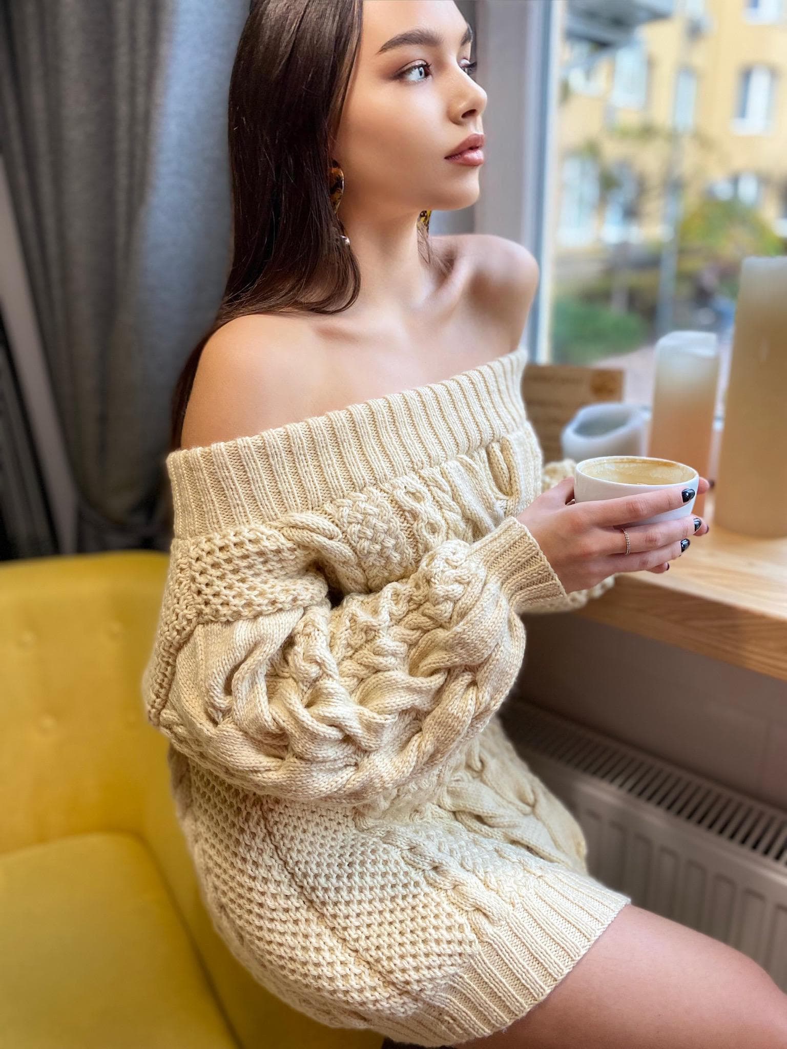 May include: A woman wearing a cream-colored off-the-shoulder cable knit sweater dress. She is sitting on a yellow chair and holding a cup of coffee.