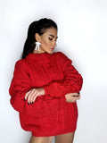 May include: A vibrant red cable-knit sweater with a high neckline. The sweater features a textured design and long sleeves. The model is wearing white bow-shaped earrings.