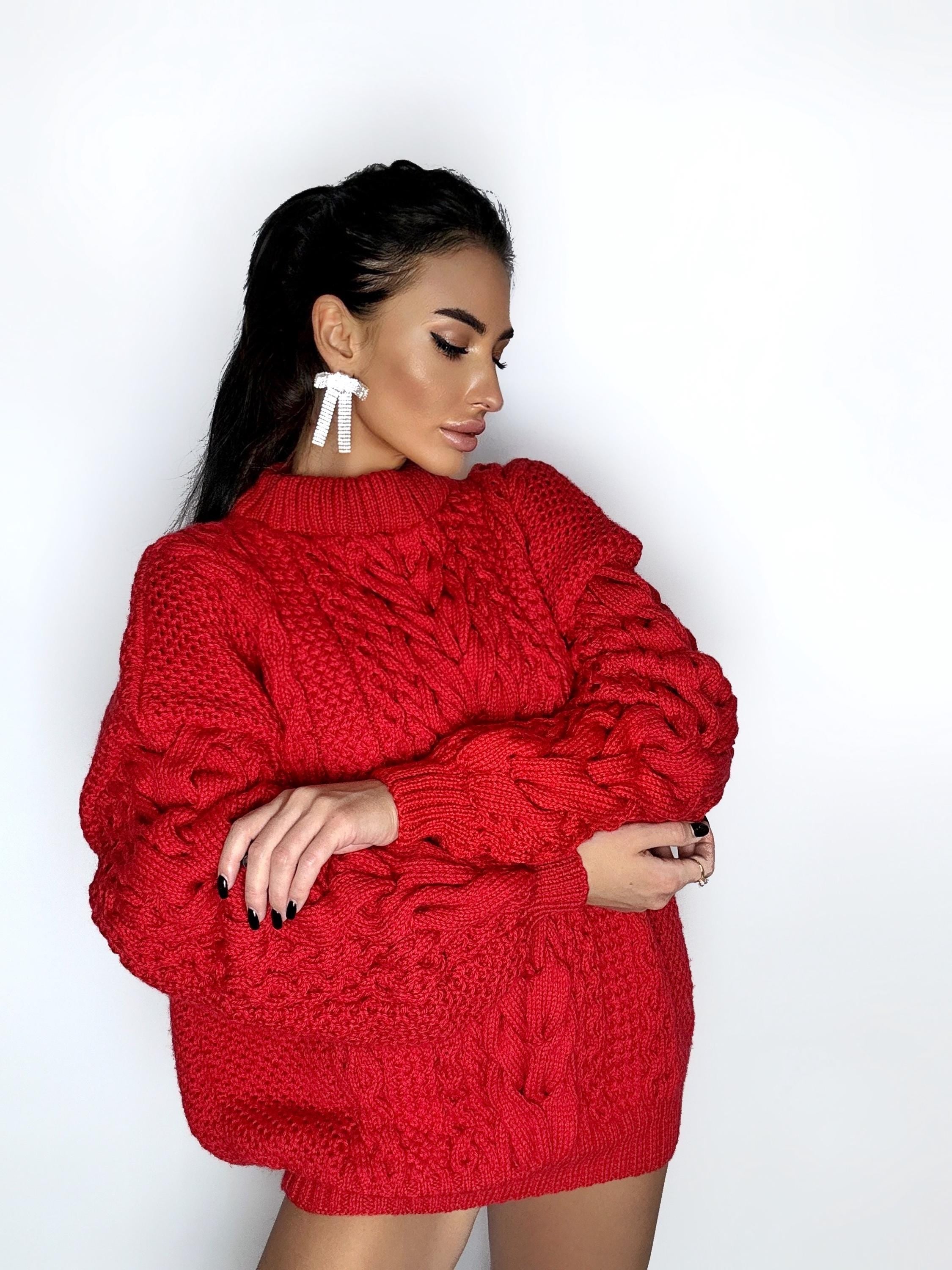 May include: A vibrant red cable-knit sweater with a high neckline. The sweater features a textured design and long sleeves. The model is wearing white bow-shaped earrings.