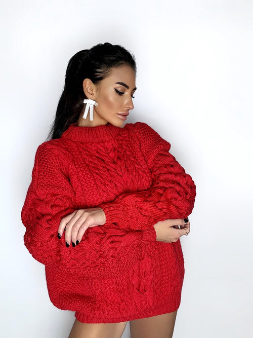 May include: A vibrant red cable-knit sweater with a high neckline. The sweater features a textured design and long sleeves. The model is wearing white bow-shaped earrings.