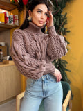 May include: A mauve-colored, chunky knit sweater with a turtleneck and cable-knit detailing. The sweater has long, voluminous sleeves and is paired with light-wash blue jeans. The overall style is casual and cozy.