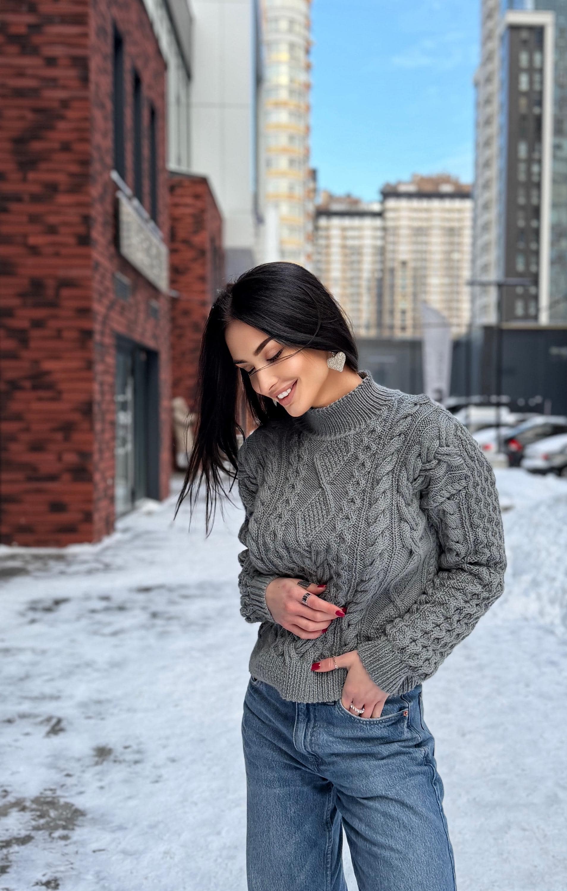 May include: A gray cable-knit sweater with a high neckline. The sweater has a textured pattern and long sleeves. The person is wearing blue jeans. The background shows a city street with buildings and snow.