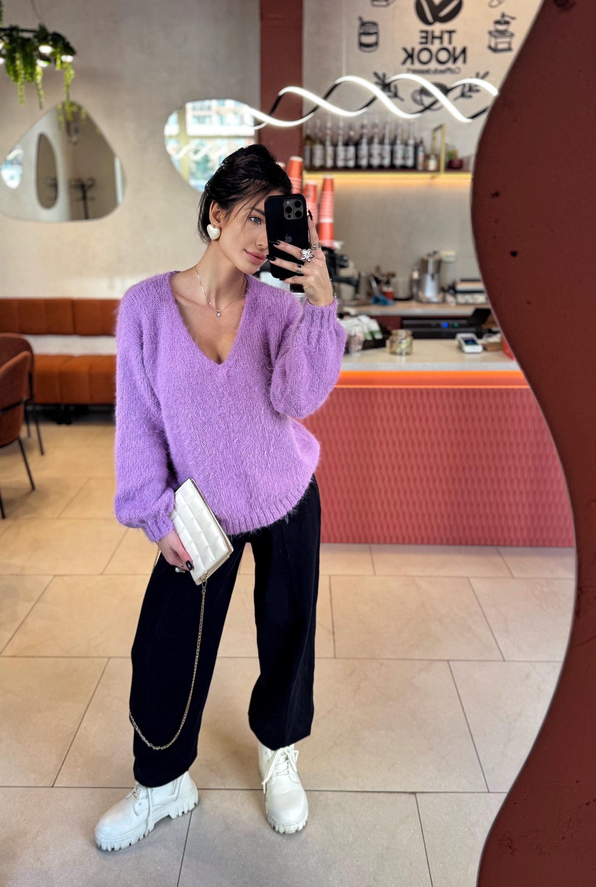 May include: A woman wearing a lilac V-neck sweater, black pants, and white boots. She is holding a white quilted clutch with a gold chain. The background includes a mirror and a cafe setting.