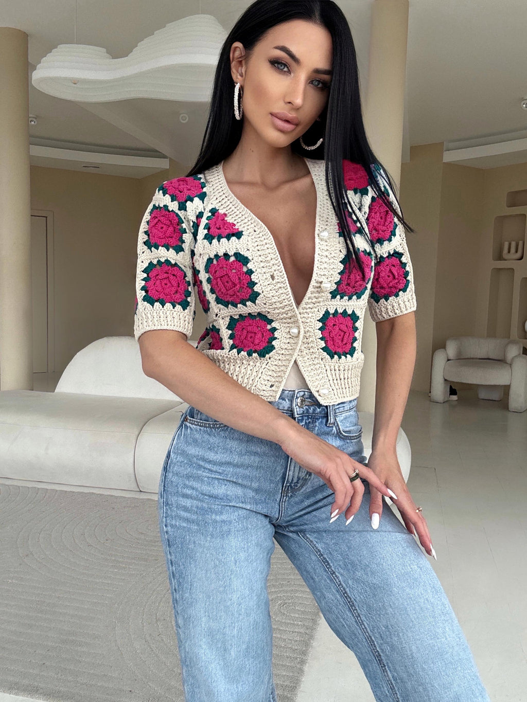 May include: Cream-colored crochet cardigan with short sleeves and a V-neck. The cardigan features a floral pattern with pink and green accents. It is buttoned up and paired with light blue jeans.