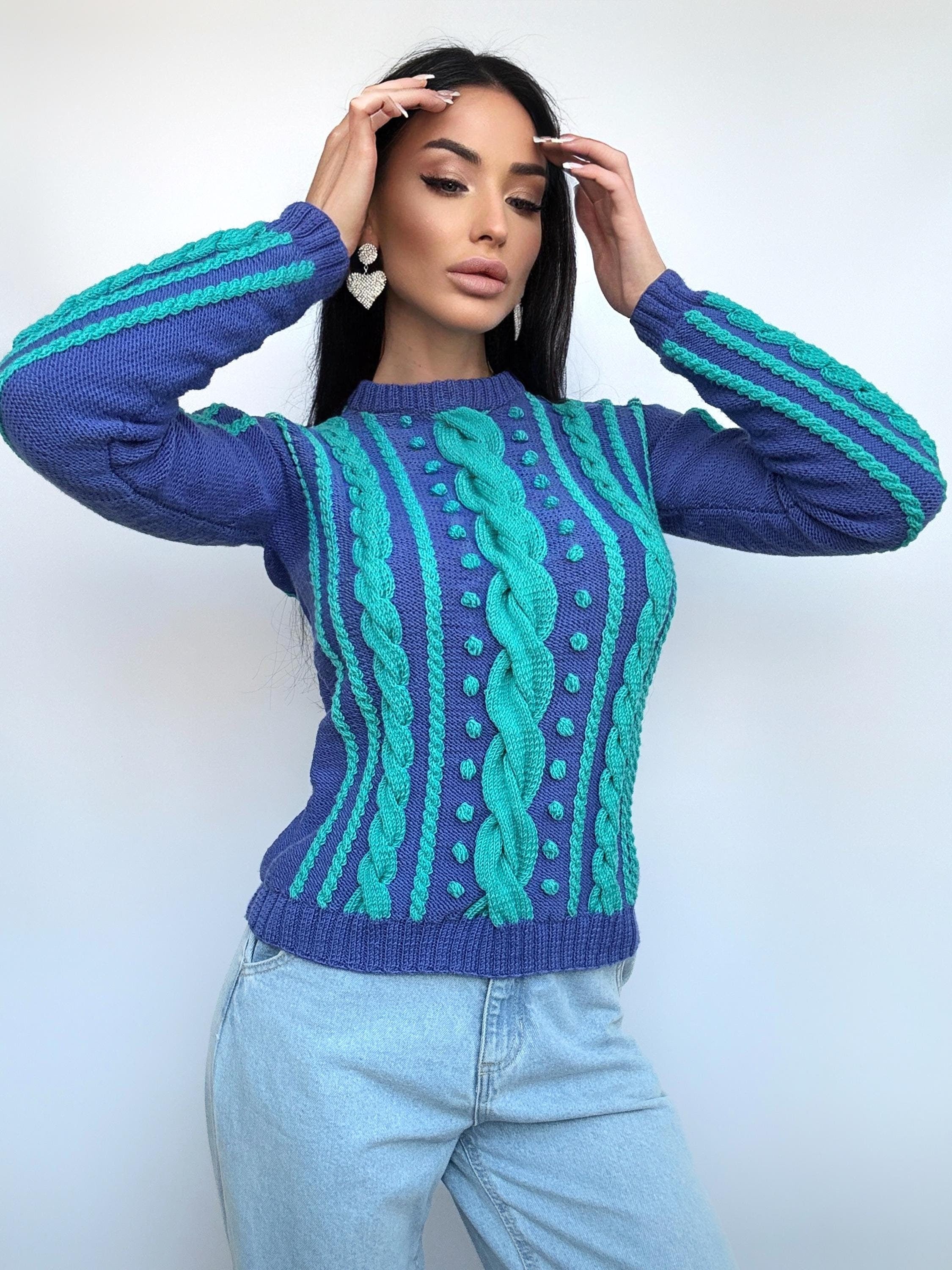 May include: A blue and teal cable-knit sweater. The sweater features a central teal cable design, teal stripes on the sleeves, and teal dotted accents. The model is wearing light blue jeans and heart-shaped earrings.