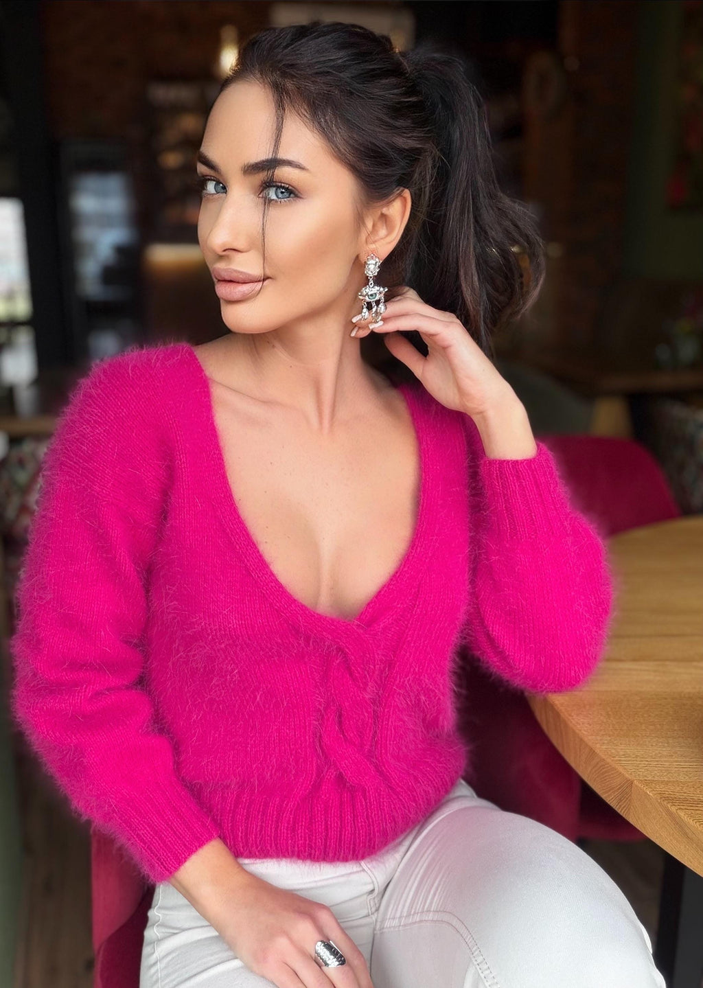 May include: A bright pink, fluffy sweater with a deep V-neck. The sweater is paired with white pants. The person is wearing silver earrings and a silver ring. The background is blurred, suggesting a restaurant setting.