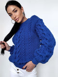 May include: A royal blue cable-knit sweater with a round neckline. The sweater features intricate cable patterns on the front and sleeves. The sleeves have a puffed design. The person is wearing white pants. The sweater is made of a soft, warm material.