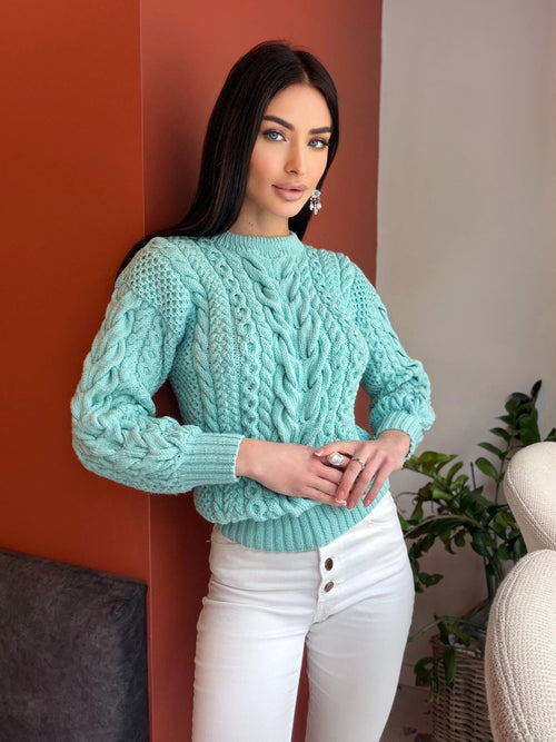 May include: A light turquoise cable-knit sweater with a crew neck and long sleeves. The sweater has a textured pattern and is paired with white pants. The model is wearing silver earrings and rings.