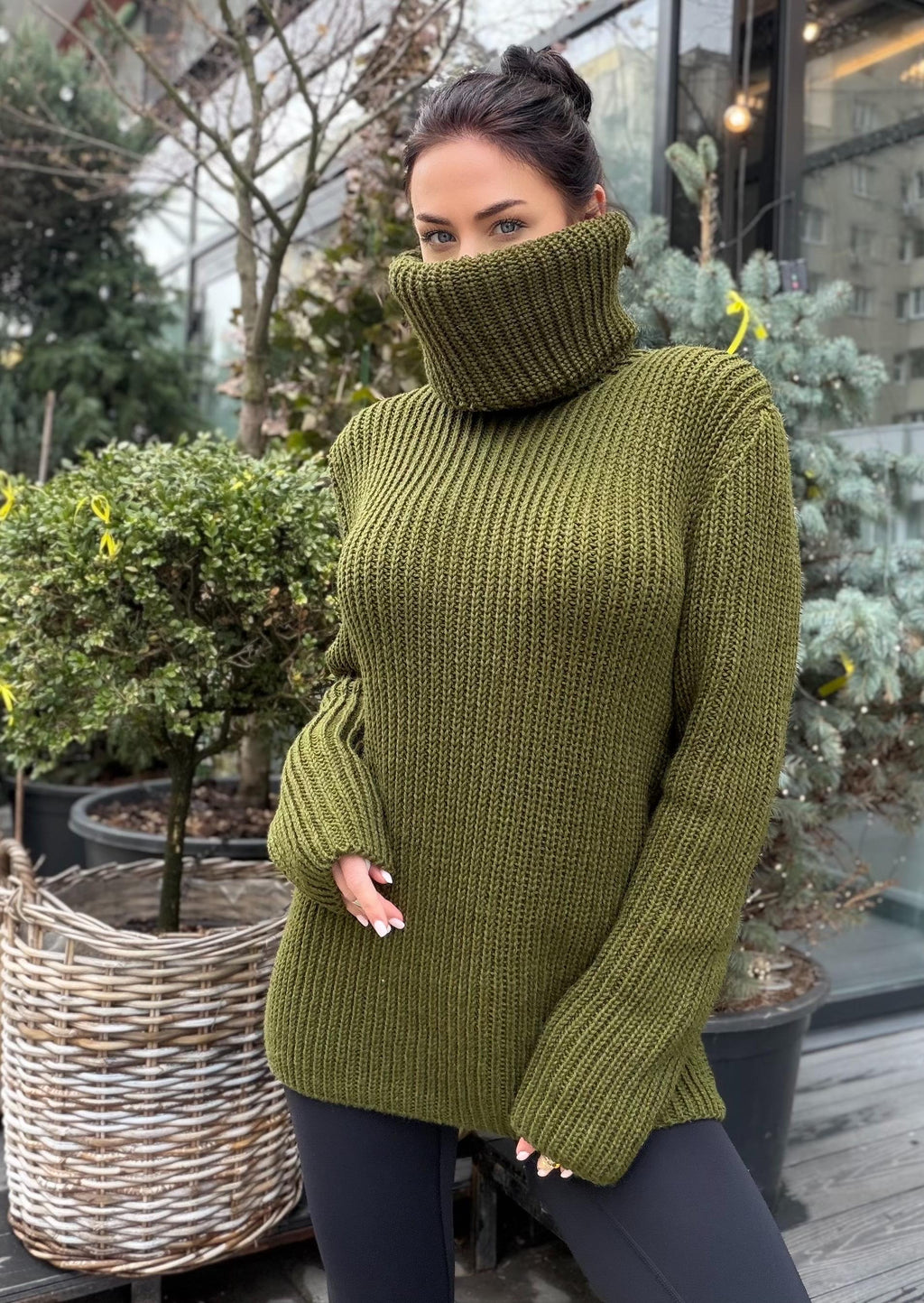 May include: Olive green turtleneck sweater with ribbed knit texture. The long-sleeved sweater has a high, wide collar and is worn with black leggings. The sweater's color is a muted, earthy tone, suitable for cooler weather.