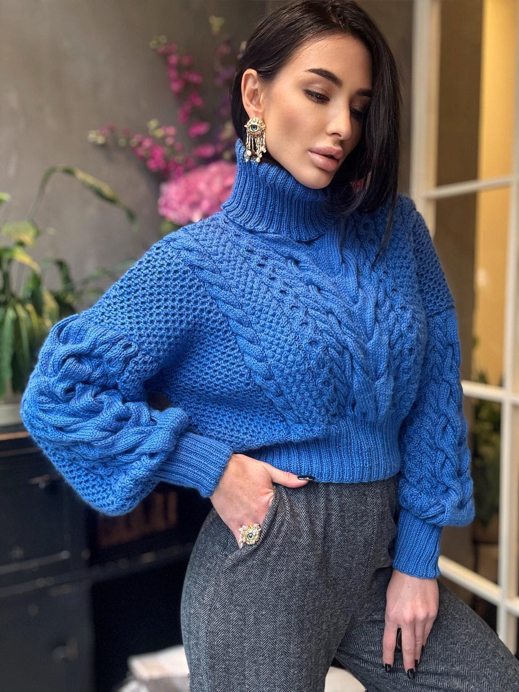 May include: A vibrant blue cable-knit sweater with a high turtleneck. The sweater features a textured pattern and long sleeves. The model is wearing gray pants and gold earrings. The sweater is a cozy and stylish garment.