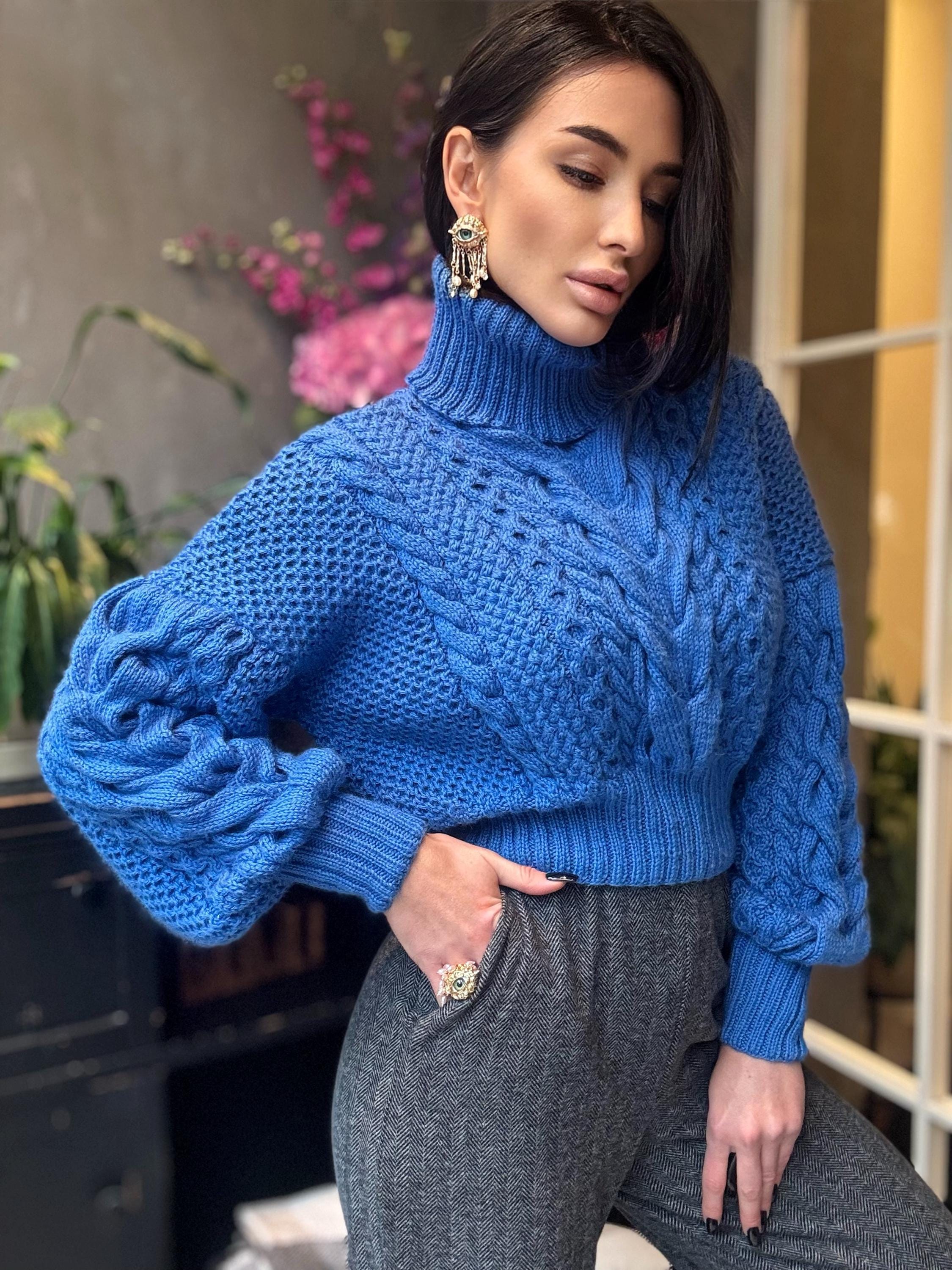 May include: A vibrant blue cable-knit sweater with a high turtleneck. The sweater features a textured pattern and long sleeves. The model is wearing gray pants and gold earrings. The sweater is a cozy and stylish garment.